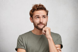 Portrait of pensive pleasant looking young male with beard and mustache, keeps hand under chin, looks thoughtfully up, tries to gather thoughts, dreams about vacations abroad in exotic country